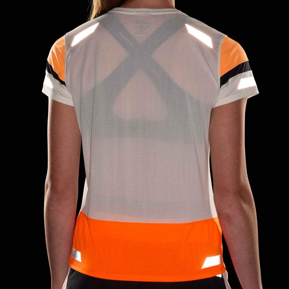 Back view of a model wearing the Brooks Women's Run Visible Short Sleeve 2.0 in the Ecru/Fluoro Flash/Black colourway. Reflective strips are reflecting artificial light. Model is also wearing Brooks shorts. (8499450806434)