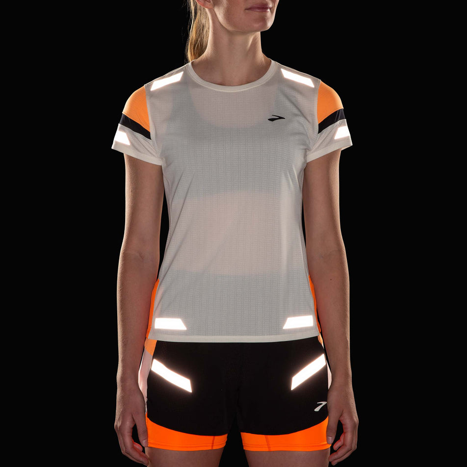 Front view of a model wearing the Brooks Women's Run Visible Short Sleeve 2.0 in the Ecru/Fluoro Flash/Black colourway. Reflective strips are reflecting artificial light. Model is also wearing Brooks shorts. (8499450806434)