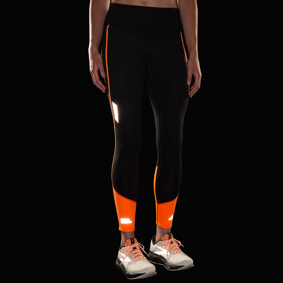 Front view of a model wearing the Brooks Women's Run Visible Tight 2.0 in the Black/Fluoro Flash colourway. Reflective strips are reflecting artificial light. Model is also wearing Brooks shoes.  (8499453362338)