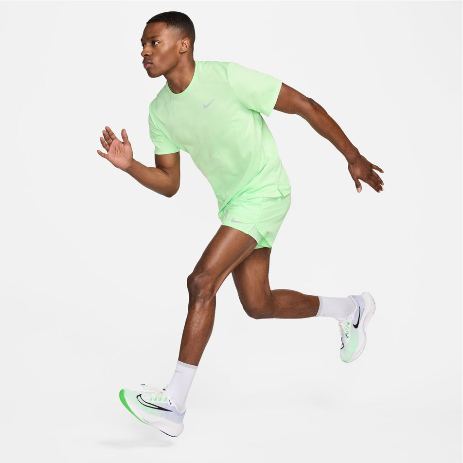 A model wearing a Nike Men's Miler Dri-FIT UV Short-Sleeve Running Top in the Vapor Green/Reflective Silv colourway. Model is also wearing Nike running shorts, socks and shoes, and is in a running pose. (8399203467426)