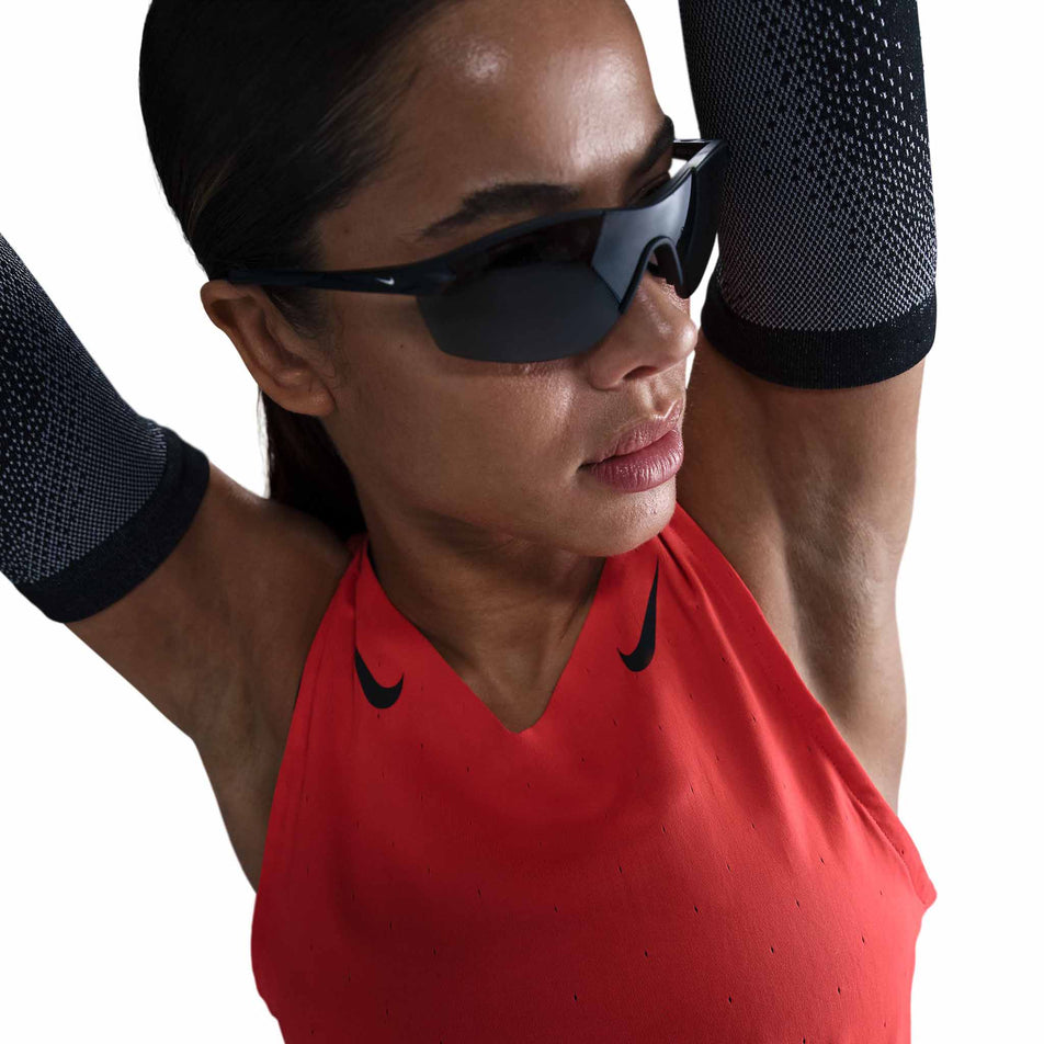 Close-up front view of a model wearing a Nike Women's AeroSwift Dri-FIT ADV Running Singlet in the Lt Crimson/Black colourway. Model is also wearing sunglasses and arm sleeves. (8562048630946)