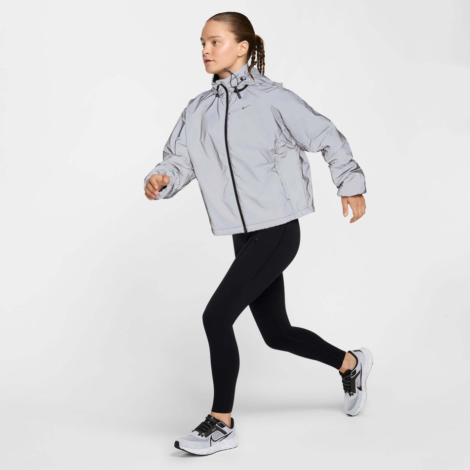 Angled view of the front of a model wearing Nike Women's Running Division Therma-FIT Reflective Running Jacket in the Black/Reflective Silv/Refblk colourway. Model is also wearing black Nike tights and Nike shoes.  (8497648730274)