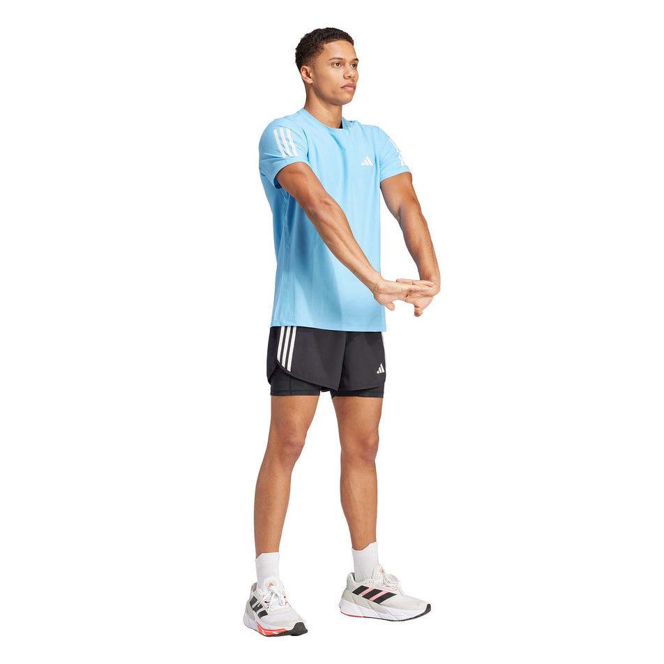Front view of a model wearing an adidas Men's Own The Run T-Shirt in the Semi Blue Burst colourway. Model is also wearing adidas shorts, shoes and socks, and is in a stretching pose. (8312074961058)