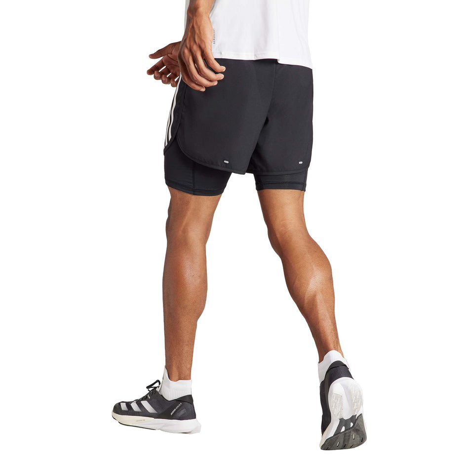 Back view of a model wearing a pair of adidas Men's Own The Run 3-Stripes 2-In-1 Shorts in the black colourway. Model is also wearing socks, shoes and a top. (8449469448354)