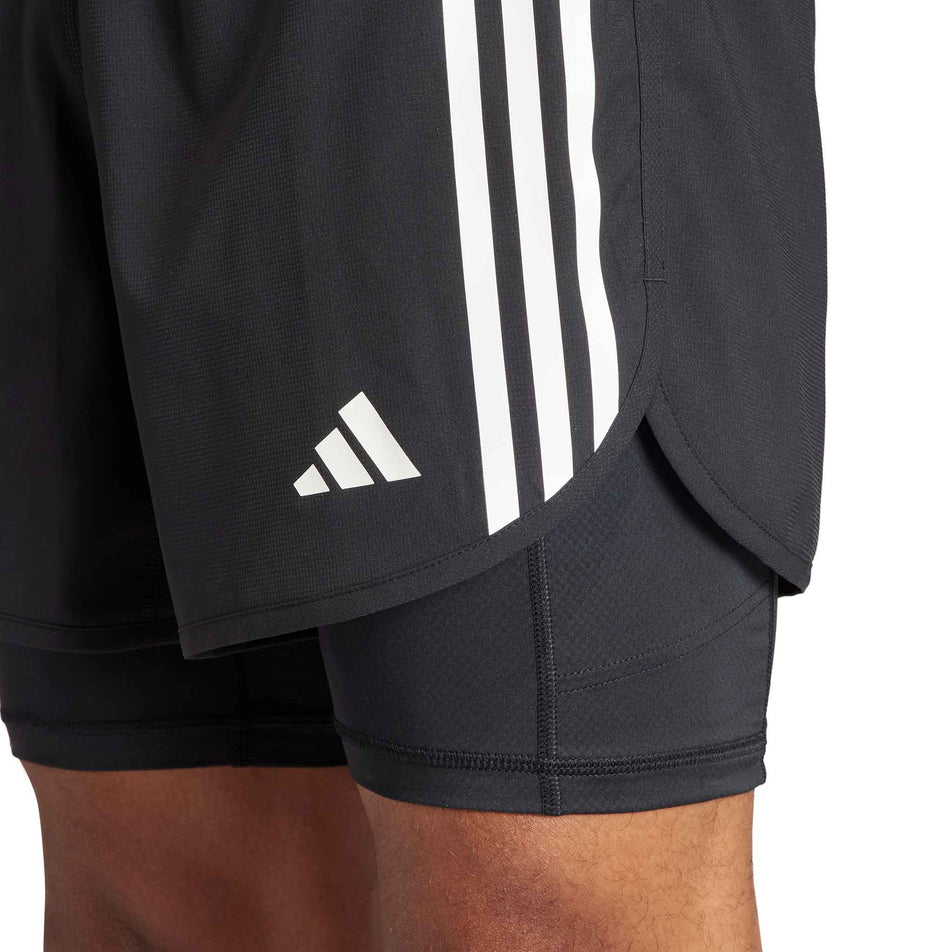 Close-up of the lower left-side a pair of adidas Men's Own The Run 3-Stripes 2-In-1 Shorts in the black colourway. Shorts are being worn by a model. (8449469448354)