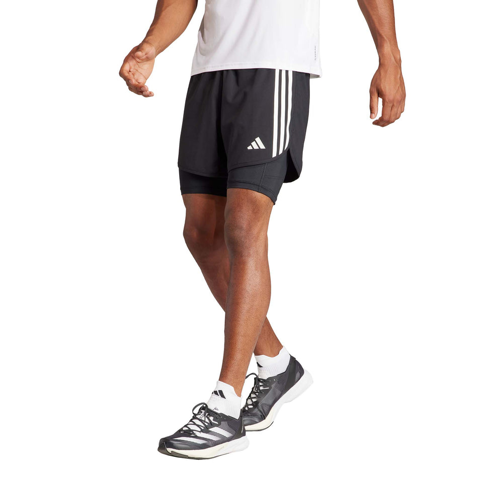 Front view of a model wearing a pair of adidas Men's Own The Run 3-Stripes 2-In-1 Shorts in the black colourway. Model is also wearing socks, shoes and a top. (8449469448354)