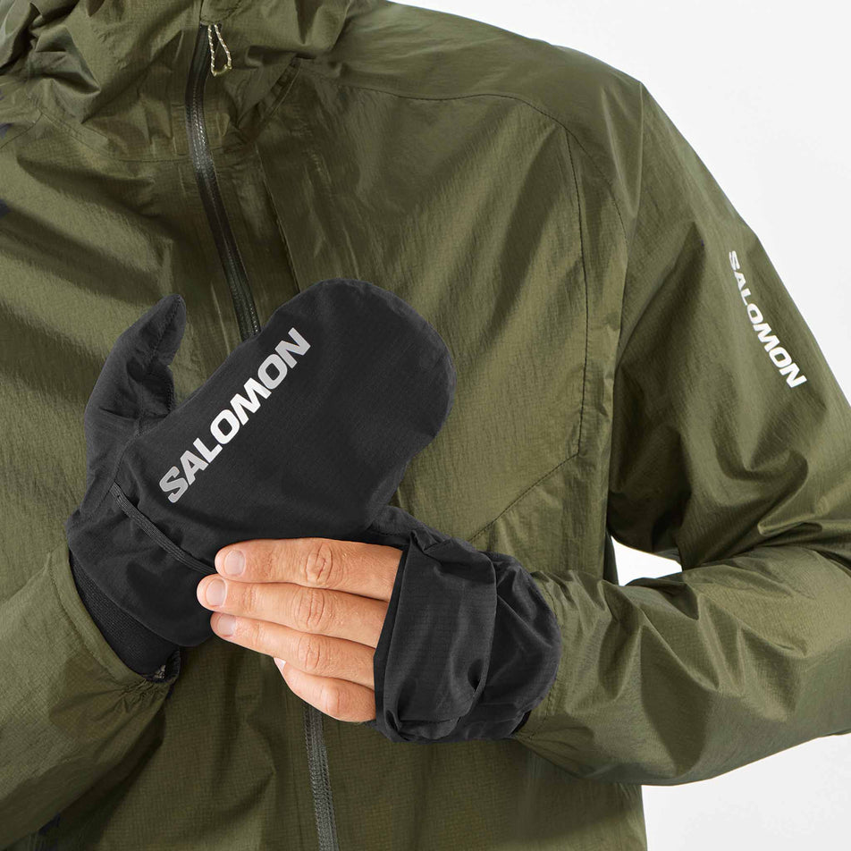 A model wearing a pair of Salomon Unisex Bonatti Waterproof Gloves in the Deep Black colourway. One glove is being worn with the mitten stored away, leaving four fingers uncovered. (8511144591522)