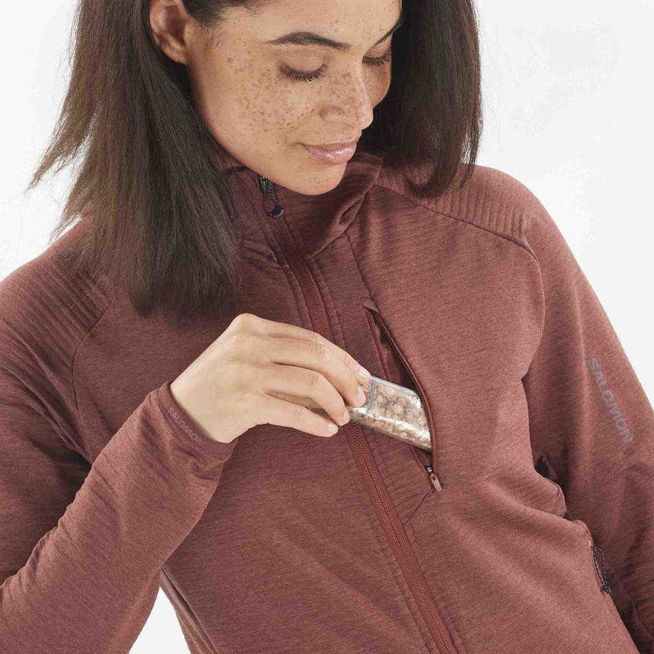 A model demonstrating that nutrition can be stored in the chest pocket of the Salomon Women's Essential Lightwarm Midlayer Jacket Hoodie. Top is being worn by a model.  (8509527097506)