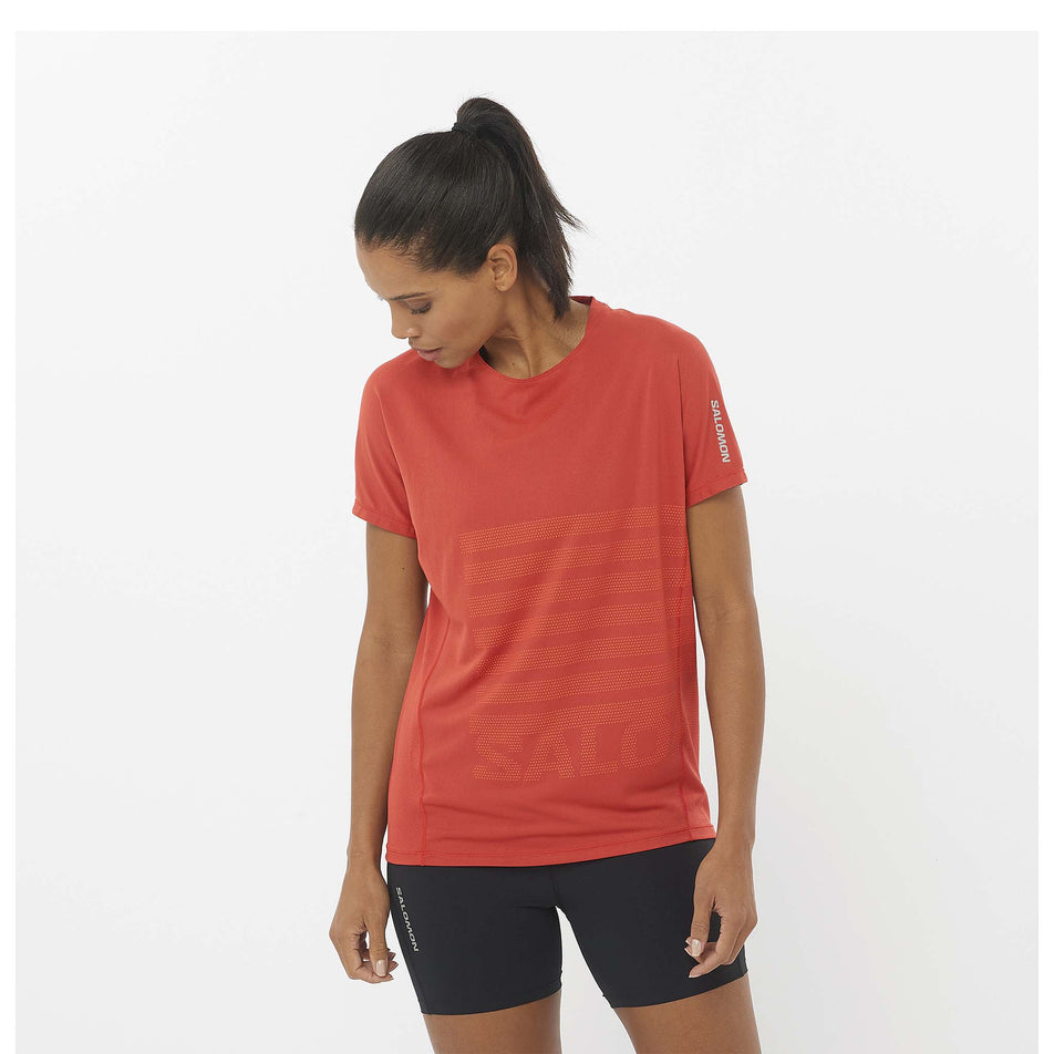 Front view of a model wearing the Salomon Women's Sense Aero Graphic Short Sleeve T-Shirt in the Tandoori Spice/Shocking Orange colourway. Model is also wearing shorts. (8464135618722)