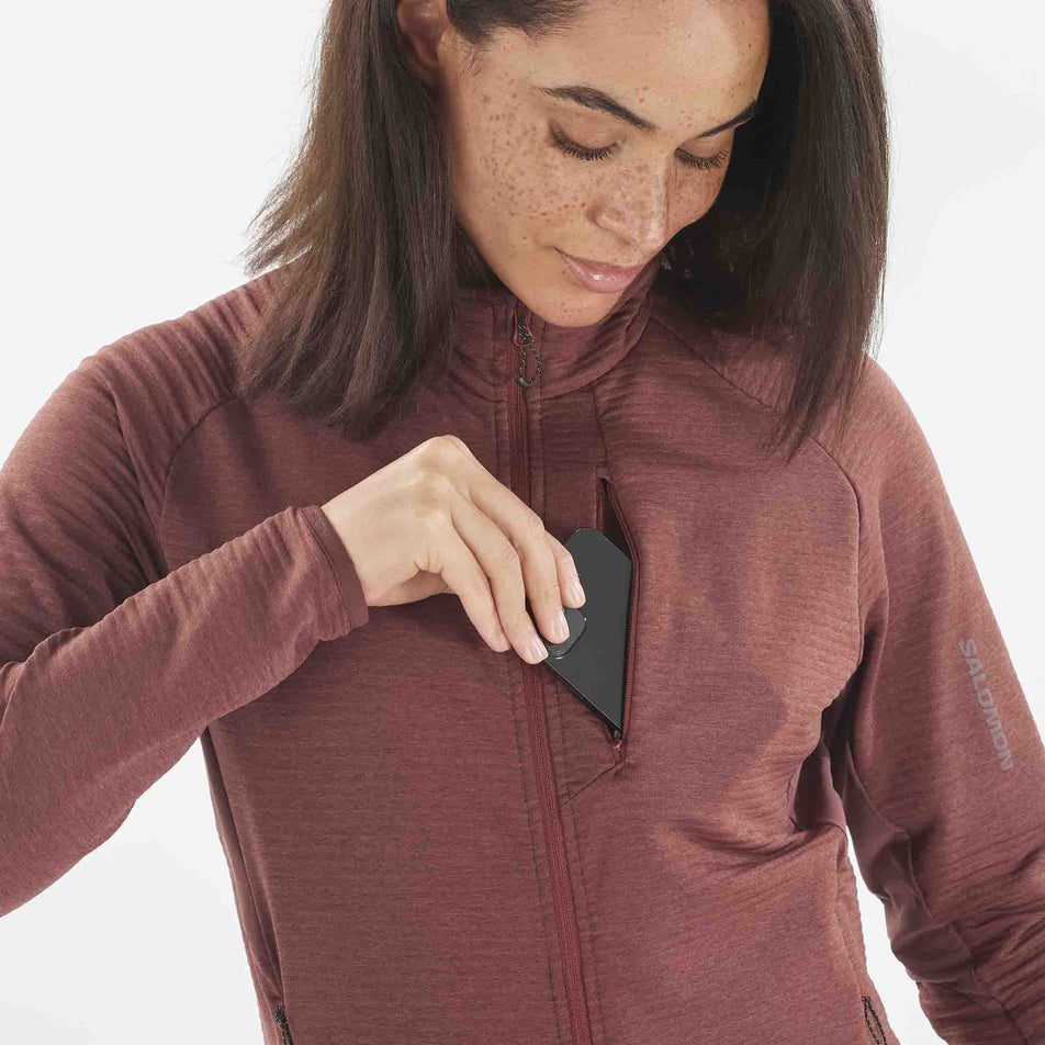 A model demonstrating that a phone can be stored in the chest pocket of the Salomon Women's Essential Lightwarm Full Zip Midlayer Jacket. The jacket is being worn by a model. (8509475389602)