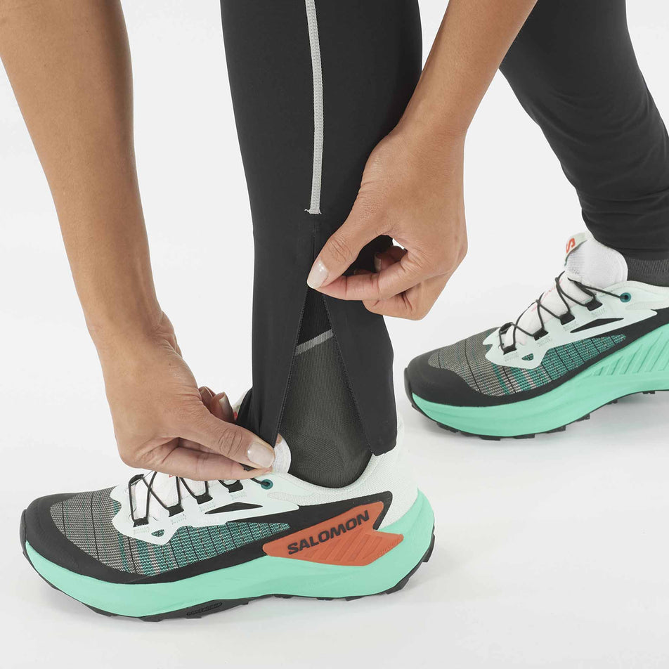 A model demonstrating the ankle zip on the left leg of the Salomon Women's Sense Aero Stow Tights. The model is wearing the tights along with running shoes. (8509442195618)