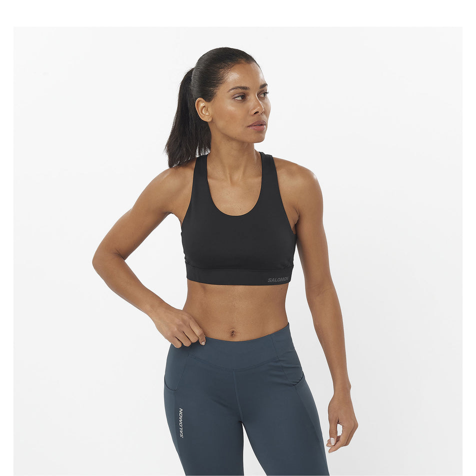 Front view of a model wearing a Salomon Women's Cross Run Sports Bra in the Deep Black colourway. Model is also wearing leggings. (8464178086050)