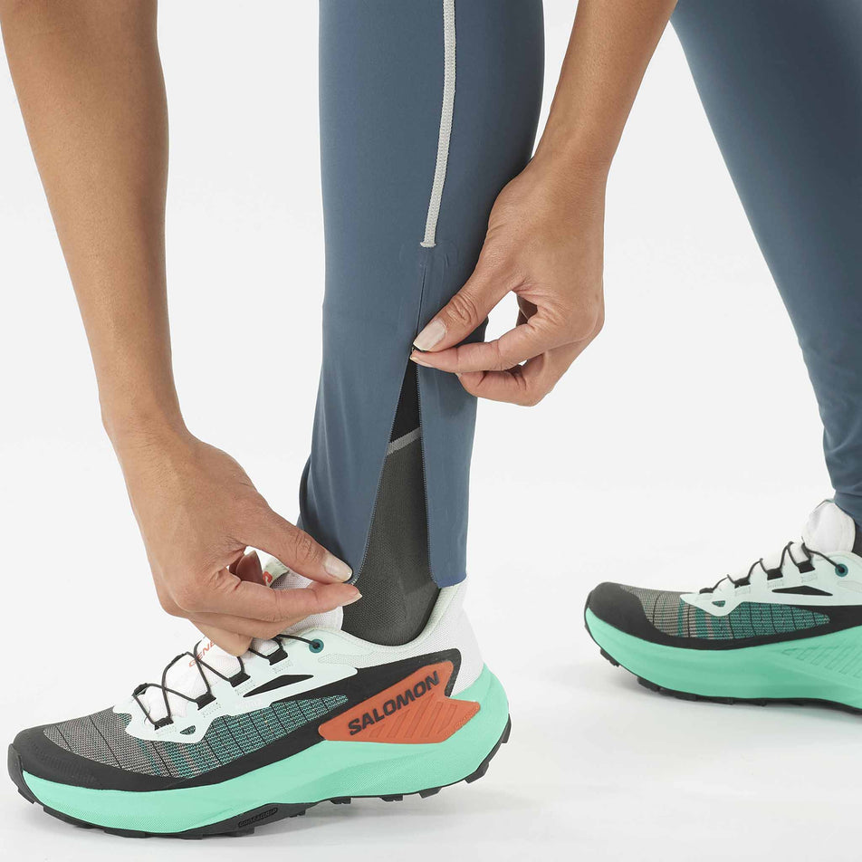 A model demonstrating the ankle zip on a pair of Salomon Women's Sense Aero Stow Tights. Model is wearing the tights and is also wearing a pair of shoes.  (8464096526498)