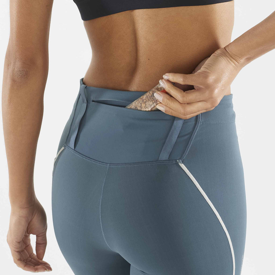 A model demonstrating that an energy bar can be stored in the zipped back pocket of a pair of Salomon Women's Sense Aero Stow Tights. Tights are being worn by a model. (8464096526498)