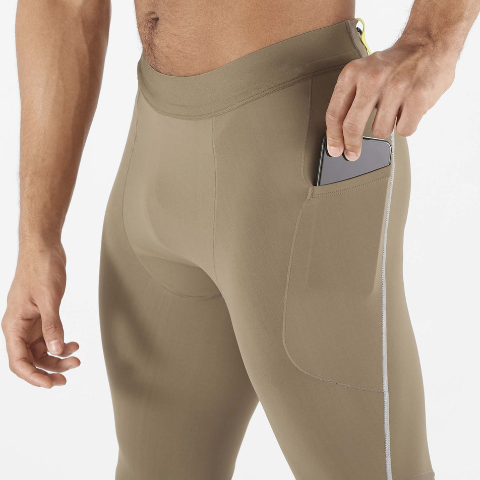 A model demonstrating that a phone can be stored on the stretch pocket on the outside of the Salomon Men's Sense Aero Stow Tights' left leg. Tights are being worn by the model. (8509599318178)